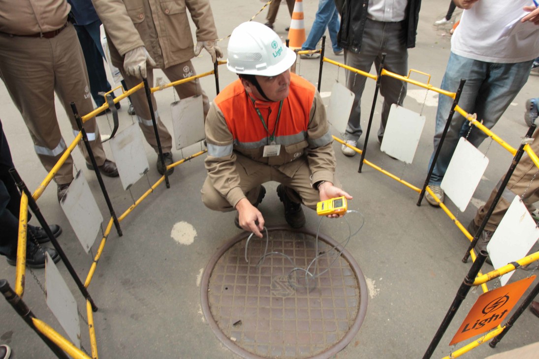 Worker in safety gear inspects a manhole cover surrounded by yellow barriers and onlookers.