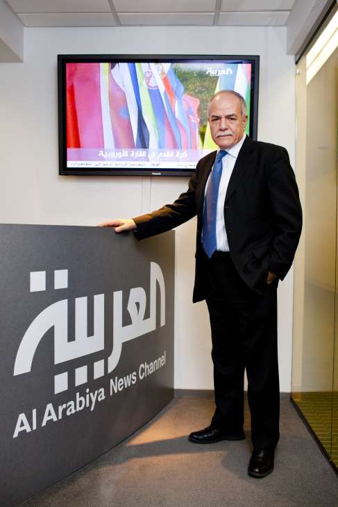 Hisham Melhem stands in Al Arabiya's Washington bureau office next to the channel's logo, with a monitor displaying Arabic news content behind him.