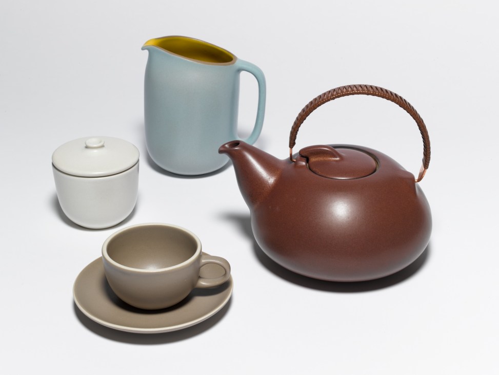 Heath Ceramics tableware collection including a white lidded container, light blue pitcher, brown teapot with woven handle, and beige cup and saucer.
