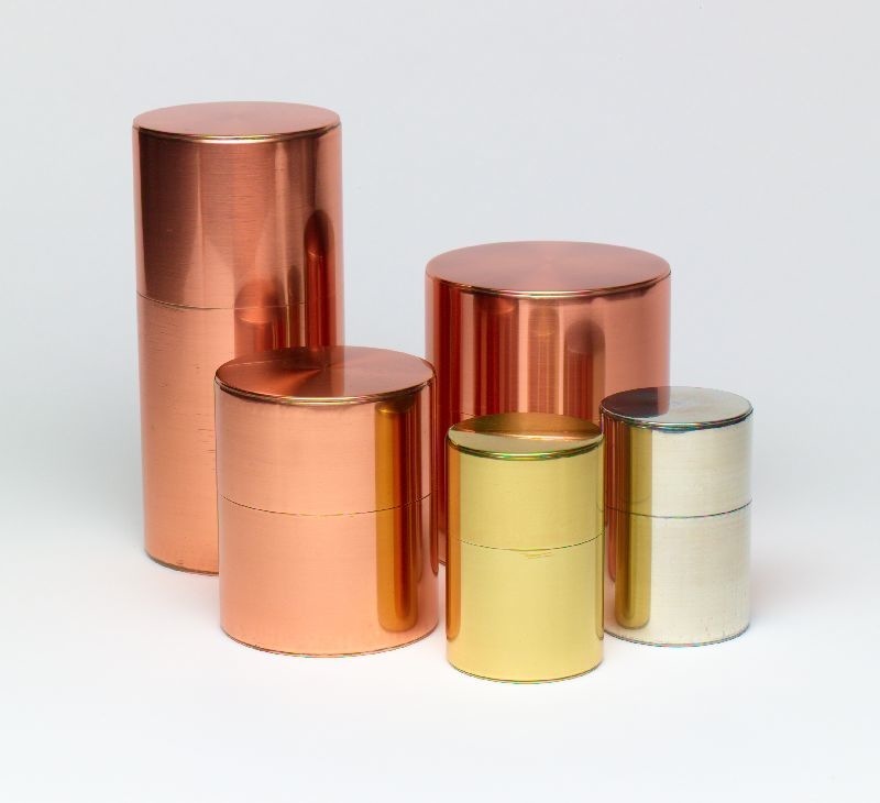 Five cylindrical metal tea caddies in copper, brass and silver finishes arranged in a group.