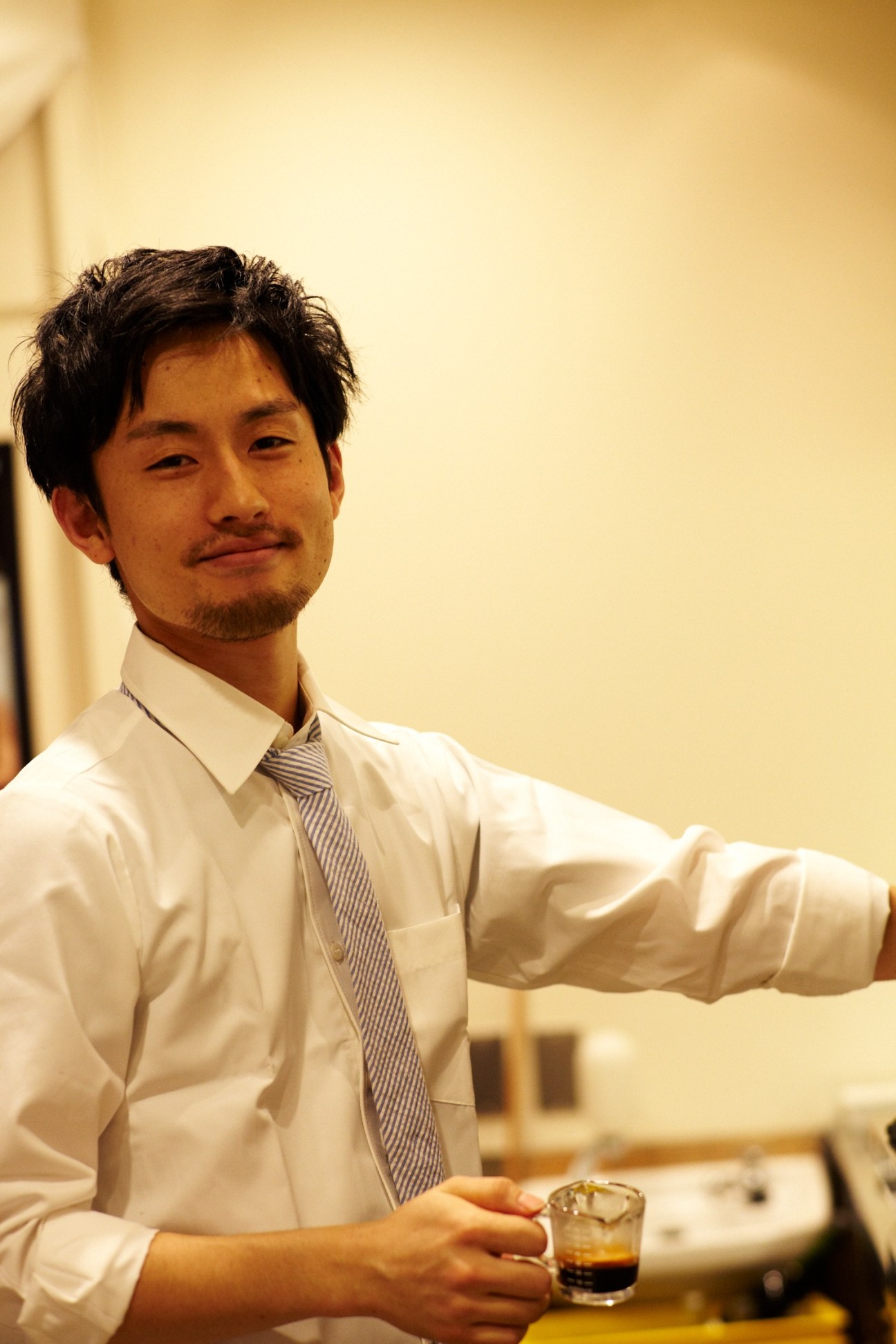 Man in white shirt and striped tie holding a glass of coffee in a café setting.