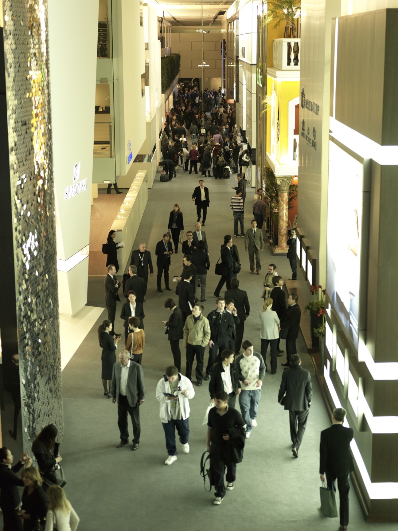 Overhead view of visitors walking through a wide corridor lined with illuminated brand displays at BaselWorld.