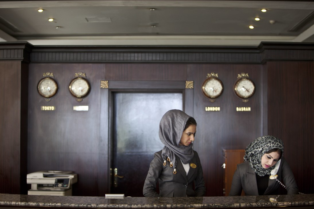 Two women in headscarves stand at the reception desk of Basra International Hotel, with world clocks displaying times for Tokyo, London and Basrah on the wall behind them.