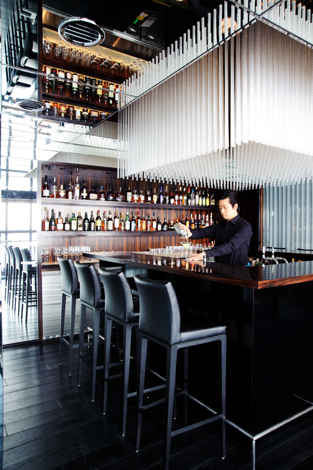 A bartender pours a drink behind a modern bar with illuminated shelving displaying bottles, featuring vertical white slats and black bar seating.