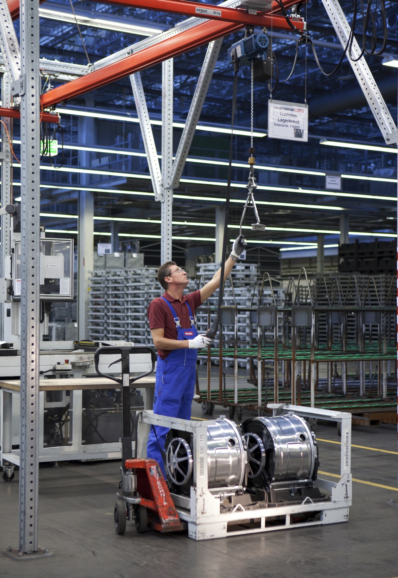 Factory worker lifting washing machine drums onto an overhead conveyor system at Miele's Gütersloh facility.