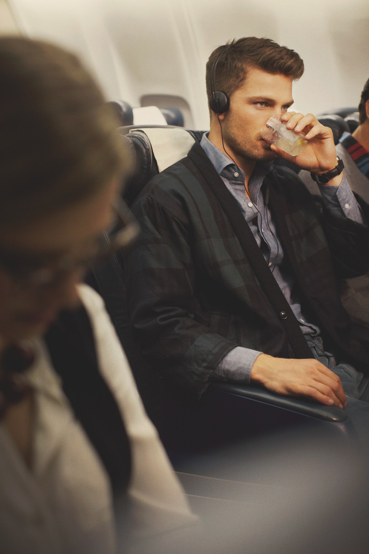 Man in airplane seat wearing headphones, jacket by ts(s), drinking from a cup