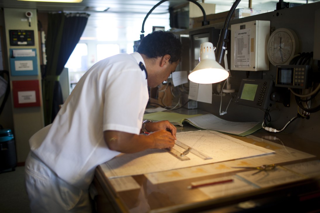 Ship officer in white uniform mapping out the day's journey on chart in control room.