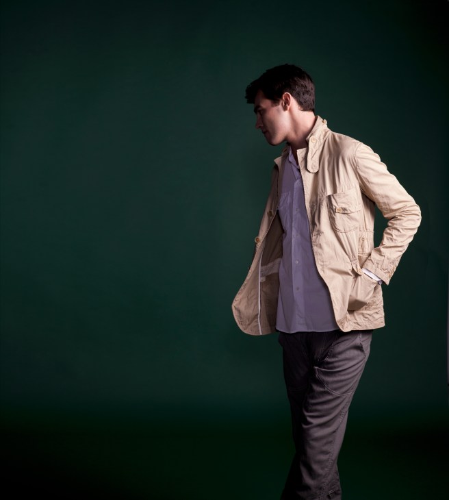 Model wearing a beige Woolrich Woolen Mills jacket over a purple shirt and dark trousers against a green background.
