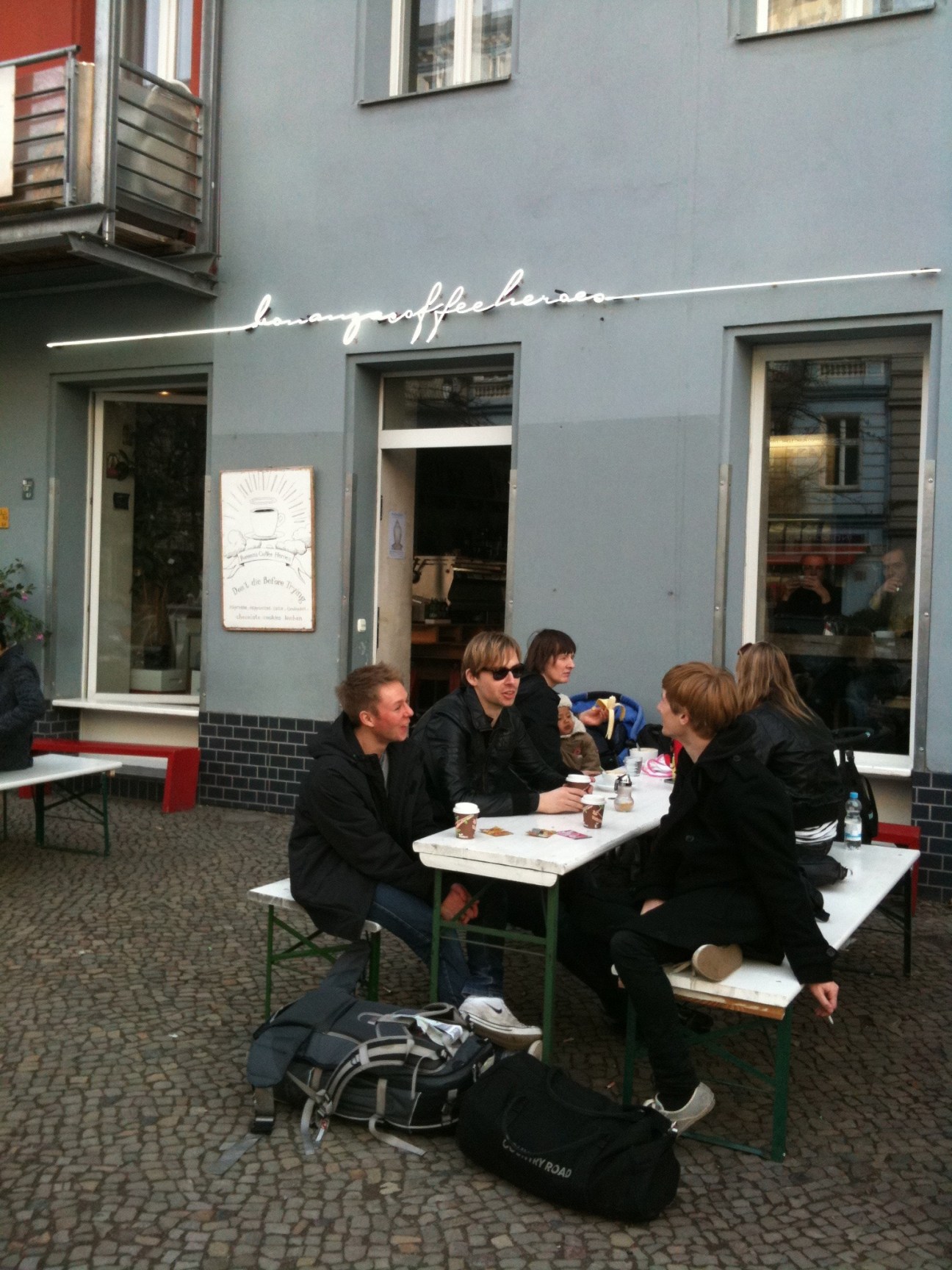 People seated at outdoor tables at Bonanza Coffee Heroes in Berlin with neon sign above.