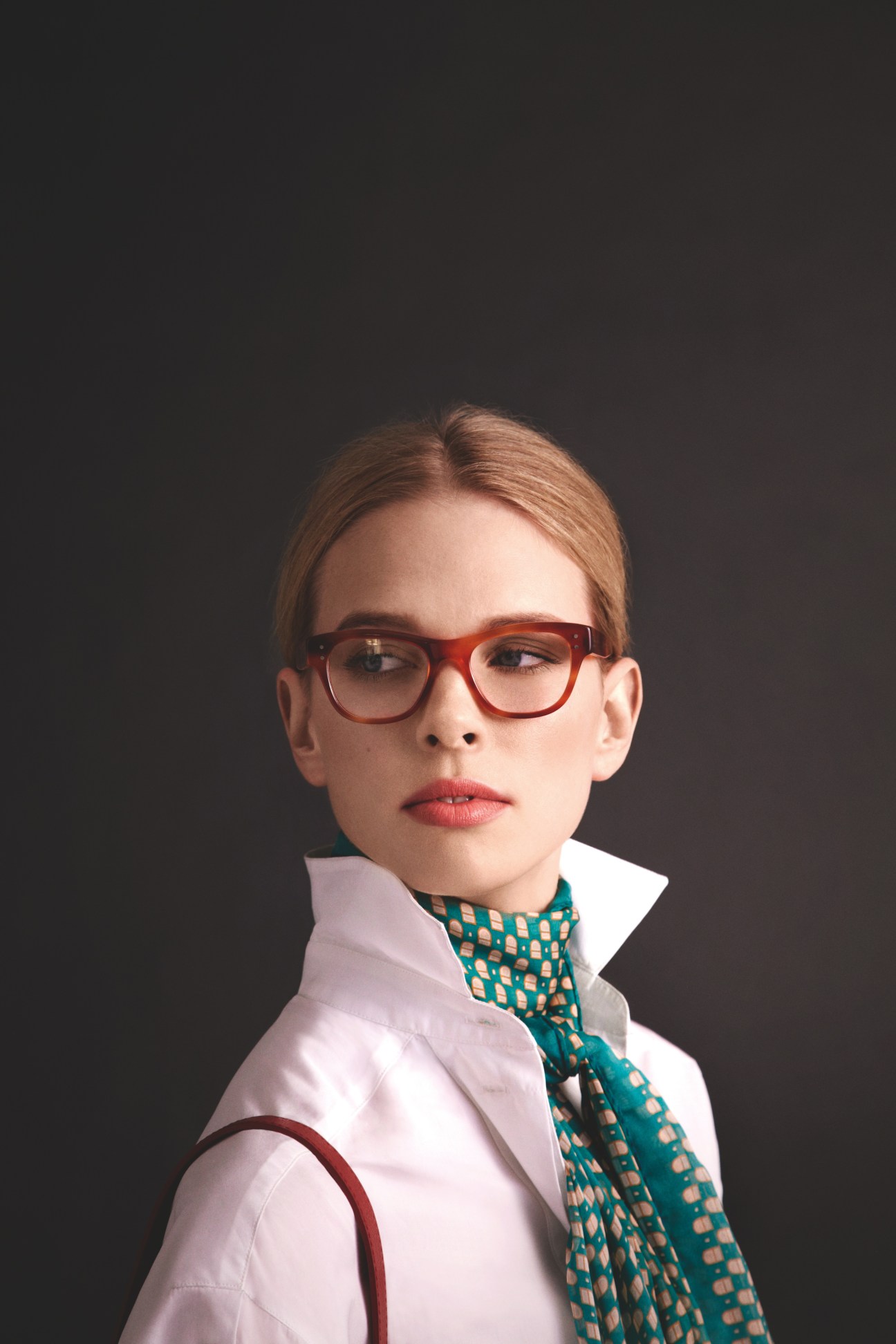Model wearing Oliver Peoples glasses, teal patterned scarf and white shirt against dark background.