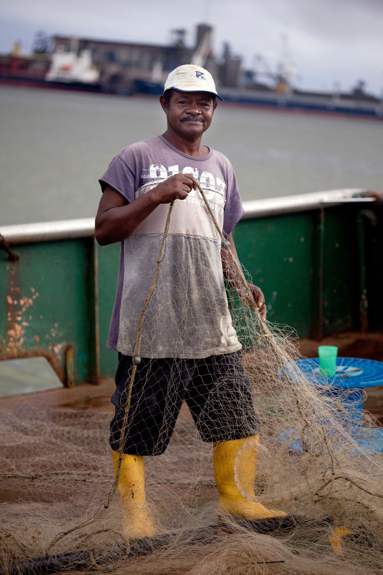 A fisherman in yellow boots holds a fishing net at Buenaventura port with cargo ships in the background.