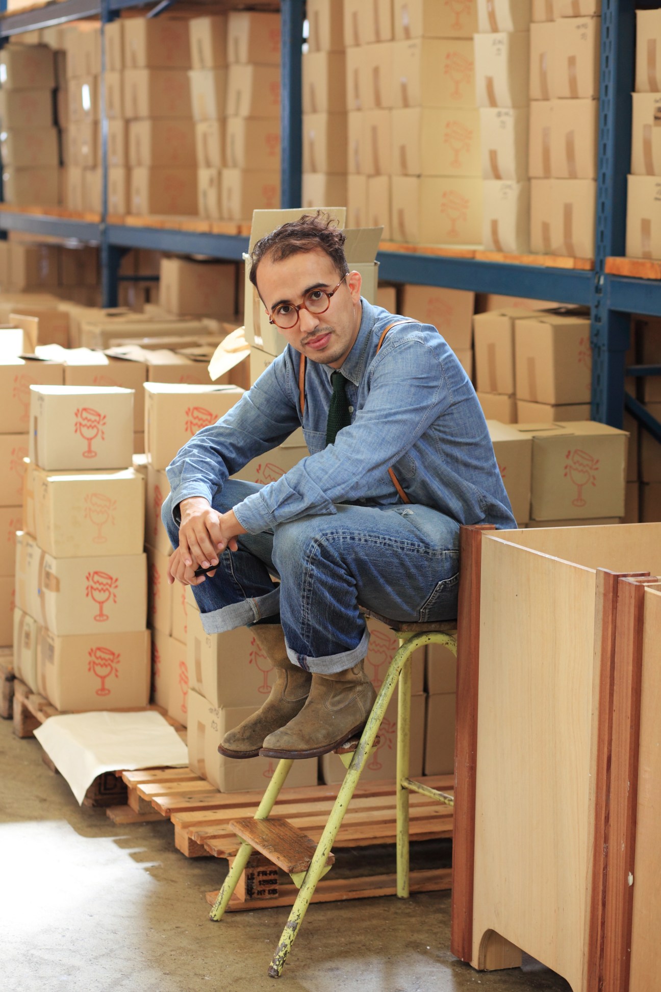 CEO Ramdane Touhami sitting on a ladder surrounded by cardboard boxes of Cire Trudon candles in a warehouse.