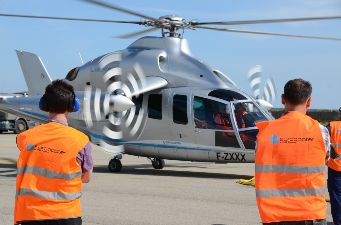 Eurocopter X3 helicopter on tarmac with orange-vested ground crew members standing nearby.