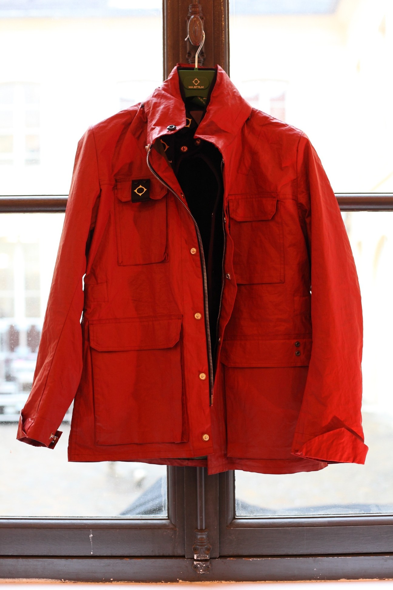 Red Ma.strum jacket with multiple chest pockets displayed on a hanger against a window.
