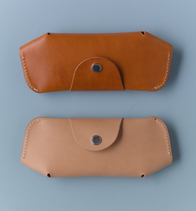 Two tan leather spectacle cases with white hand-stitched seams and metal snap buttons against a blue background.