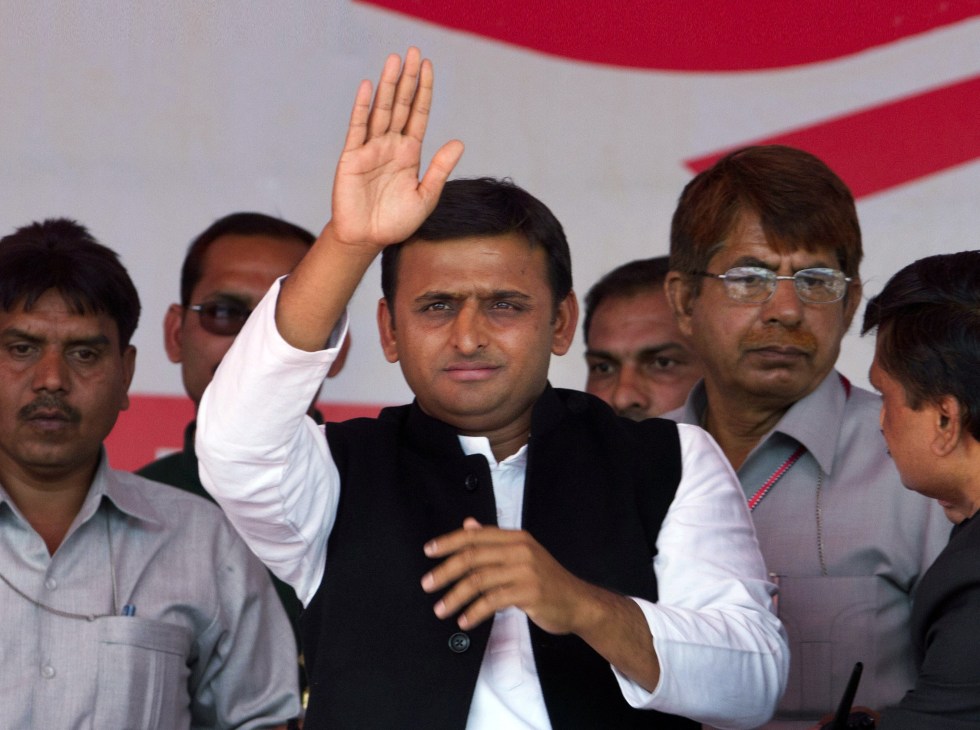 Akhilesh Yadav waving to supporters at a political event with red and white backdrop.