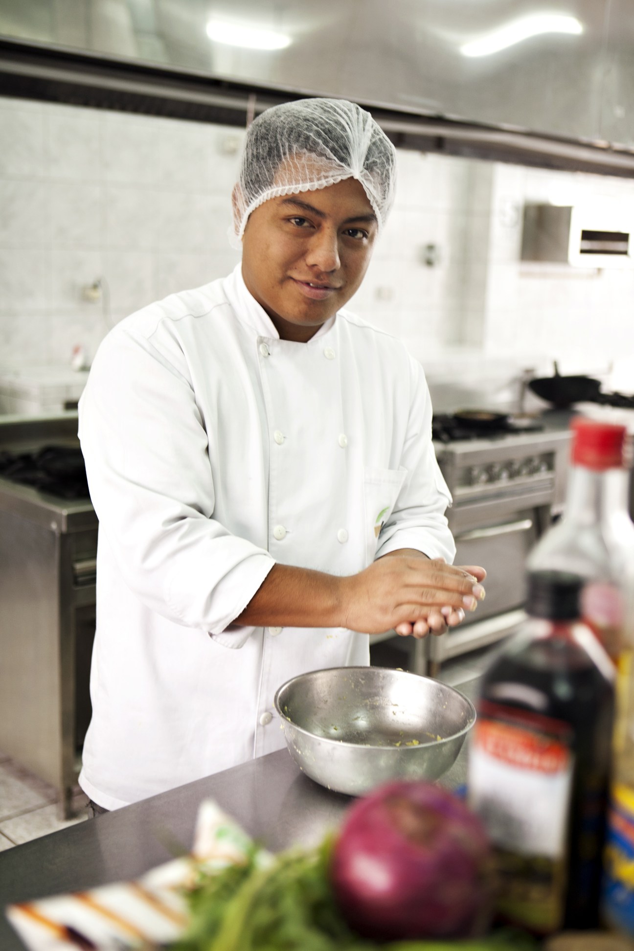 Young chef in white uniform and hair net prepares causa dish in professional kitchen.