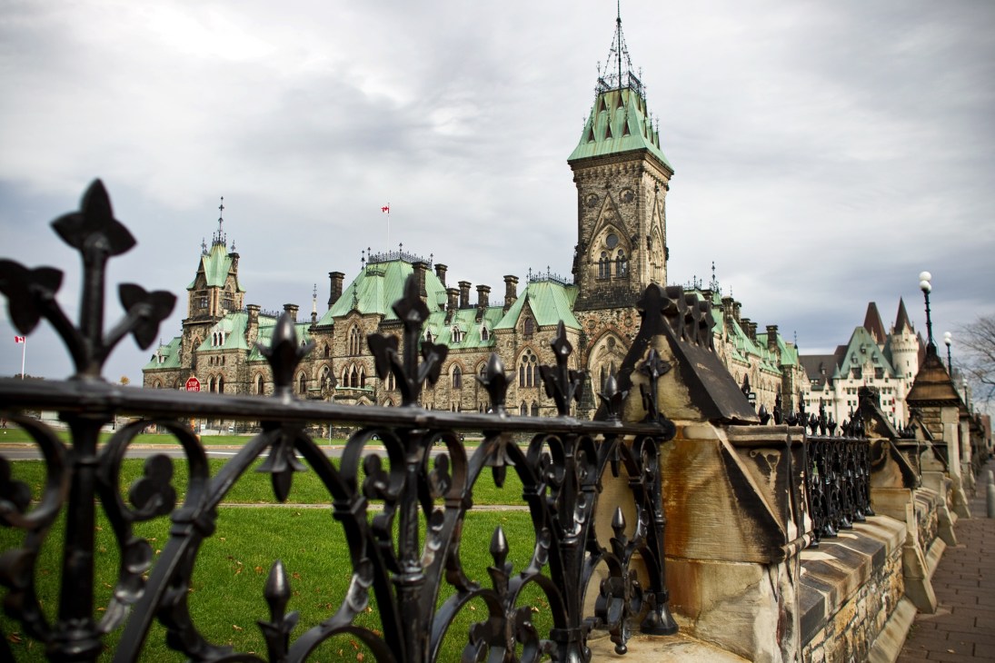 Parliament buildings in Ottawa with ornate iron fence in foreground and green-roofed Gothic Revival architecture.