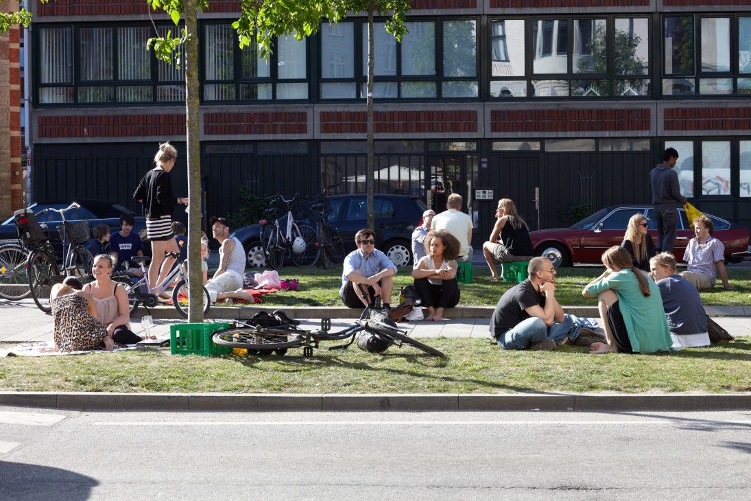 People gathering and relaxing on grass in an urban park with bicycles and modern buildings in background.
