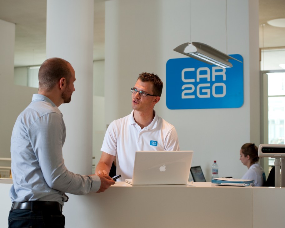Car2go staff member at reception desk with customer in modern office space with blue car2go logo on wall.
