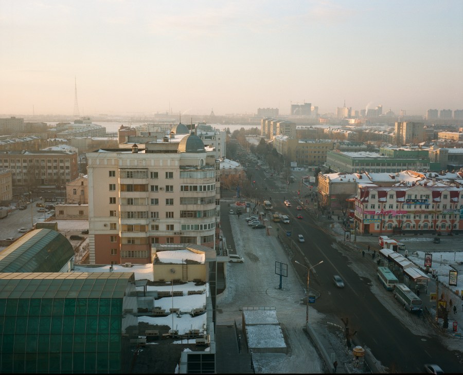 Wide aerial view of Blagoveshchensk's main street with Soviet-era apartment buildings, a frozen river visible in the distance, and hazy cityscape beyond.