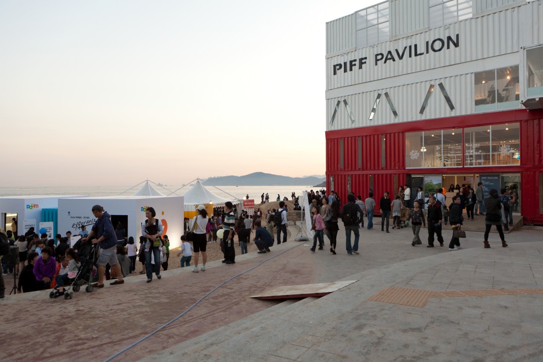 Festival visitors gather outside the PIFF Pavilion, a red and white shipping container structure on a beach with mountains visible across the water.