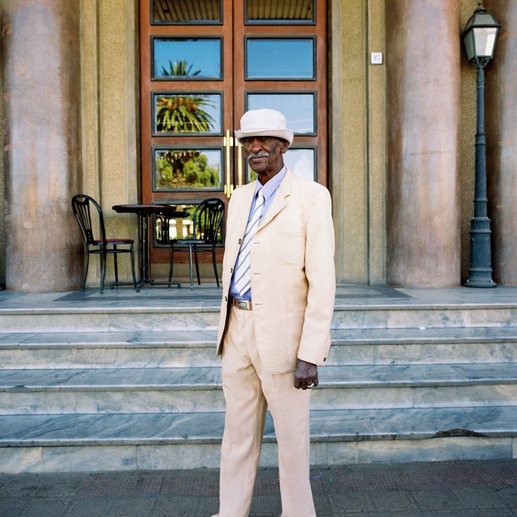 Elderly man in cream suit and white hat standing outside Italian colonial building in Asmara, Eritrea.