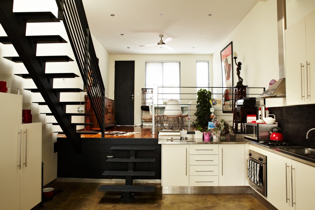 Modern kitchen and living room with cream cabinets, black countertops, and a striking black staircase with metal railing.