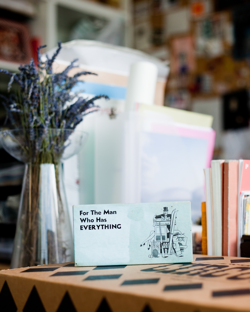 A desk detail showing a greeting card reading 'For The Man Who Has EVERYTHING' with a robot illustration, surrounded by lavender flowers and colorful papers.