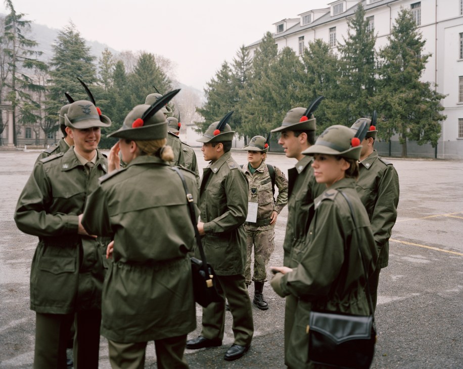 Italian Alpini soldiers in uniform with distinctive plumed hats gather in a barracks courtyard before the opening ceremony.