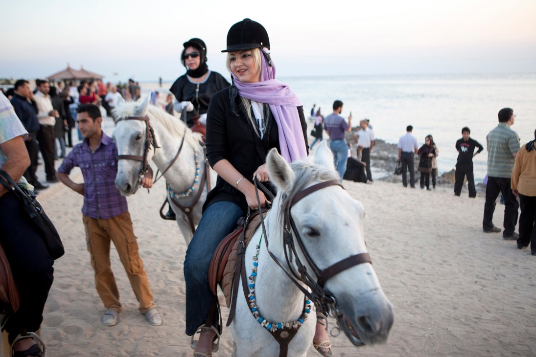 Woman in equestrian gear riding a white horse on a beach with tourists gathering at sunset on Kish island.