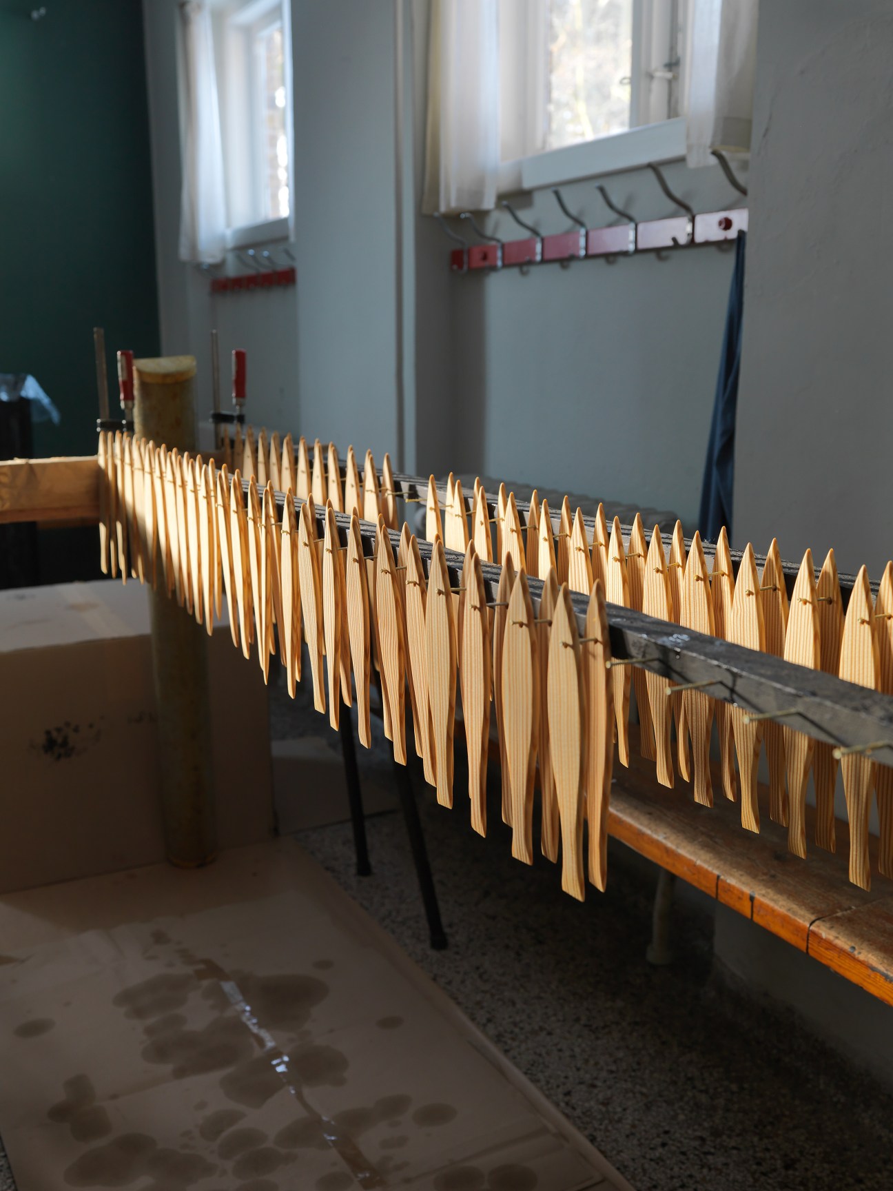 Wooden fish-shaped pieces for a Flensted mobile hanging from a drying rack in a workshop with windows.