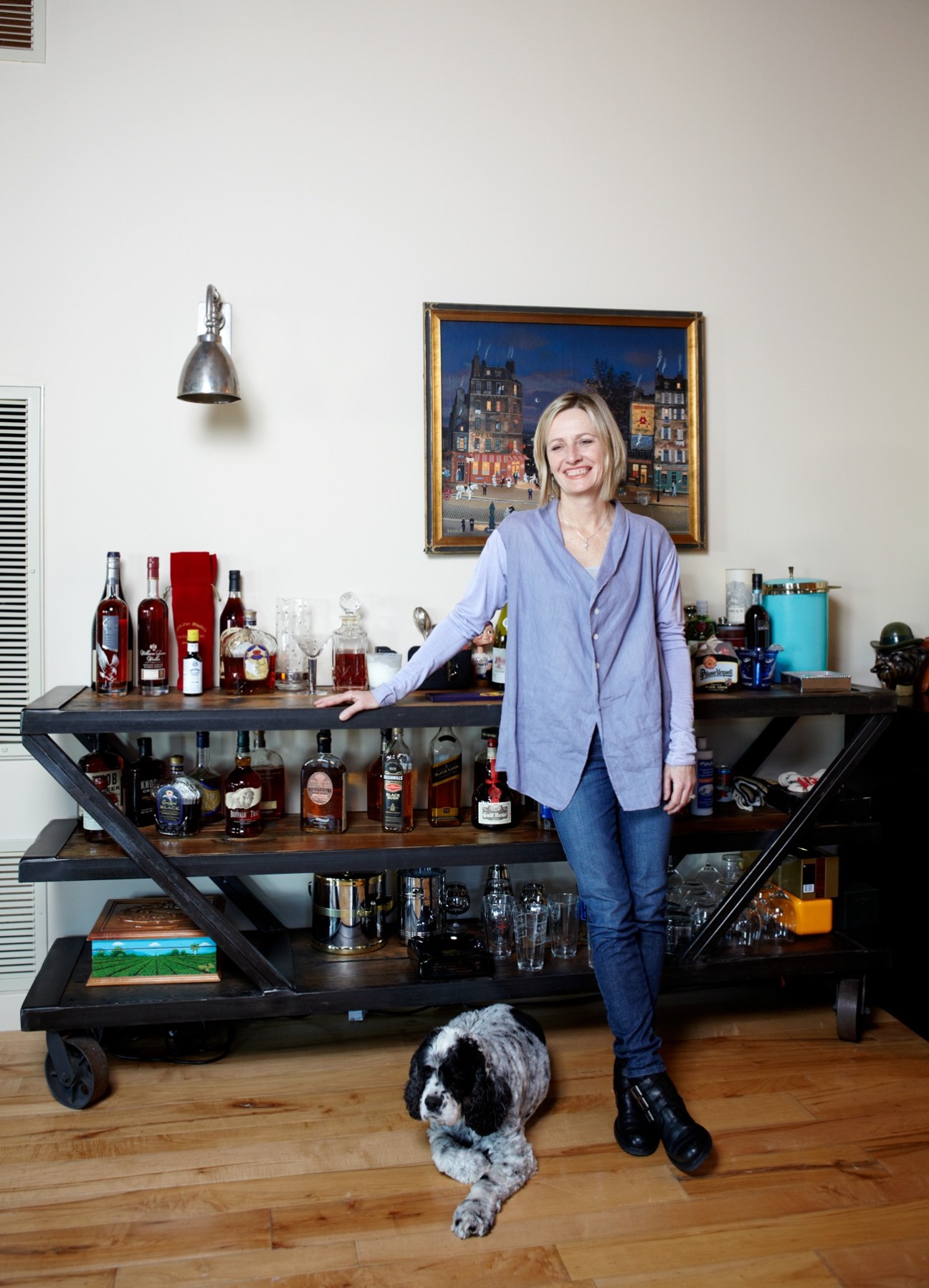 Local resident Marcy Harms stands in her loft beside a bar cart filled with liquor bottles and glassware, with her dog at her feet.