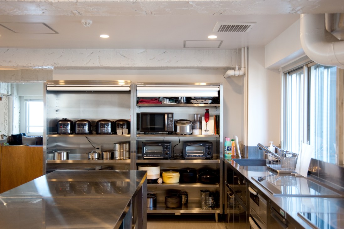 Modern shared kitchen with stainless steel counters, shelving with appliances and kitchenware, and bright windows.