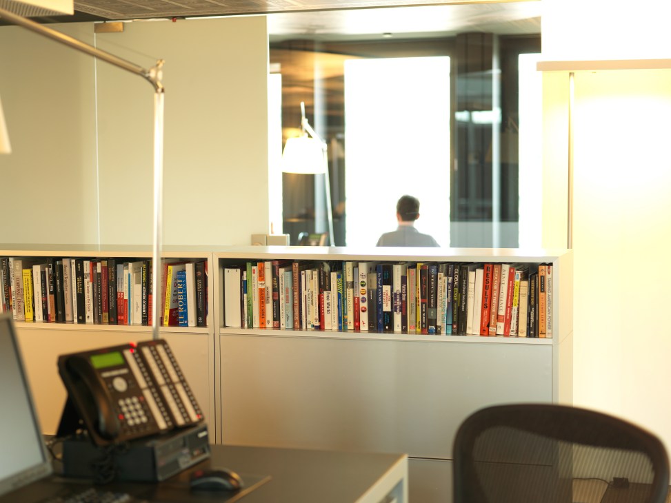 Office workspace with bookshelf, desk lamp, telephone and window view at IJ Partners Geneva.