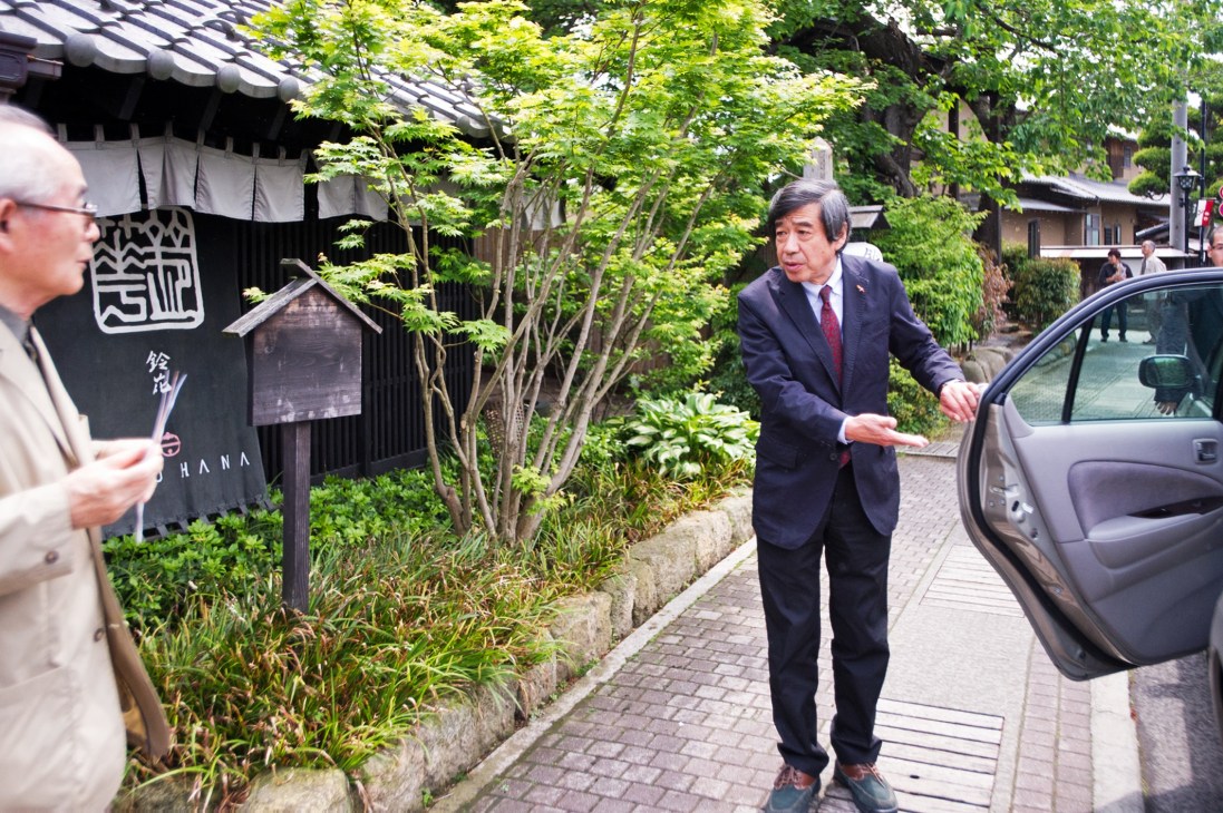 Mayor Ryozo Ichimura standing on a tree-lined street in Obuse, Japan, near a traditional building with Japanese signage.