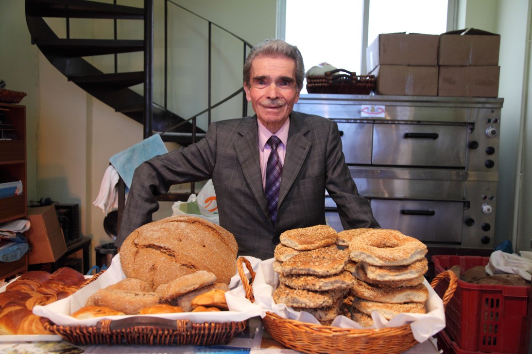 Baker Dimitri Kotsaris in a gray suit and purple tie stands behind baskets of sourdough bread and pastries in his bakery kitchen.