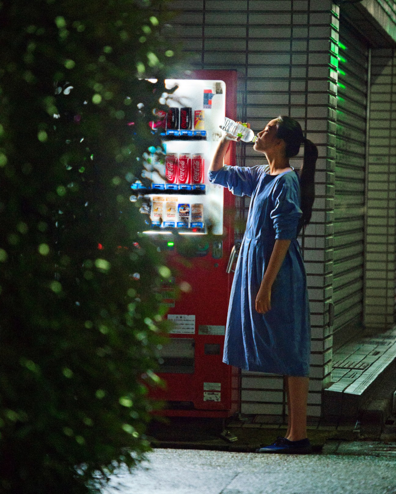 Woman in blue dress drinks from a bottle while standing at a red vending machine at night.