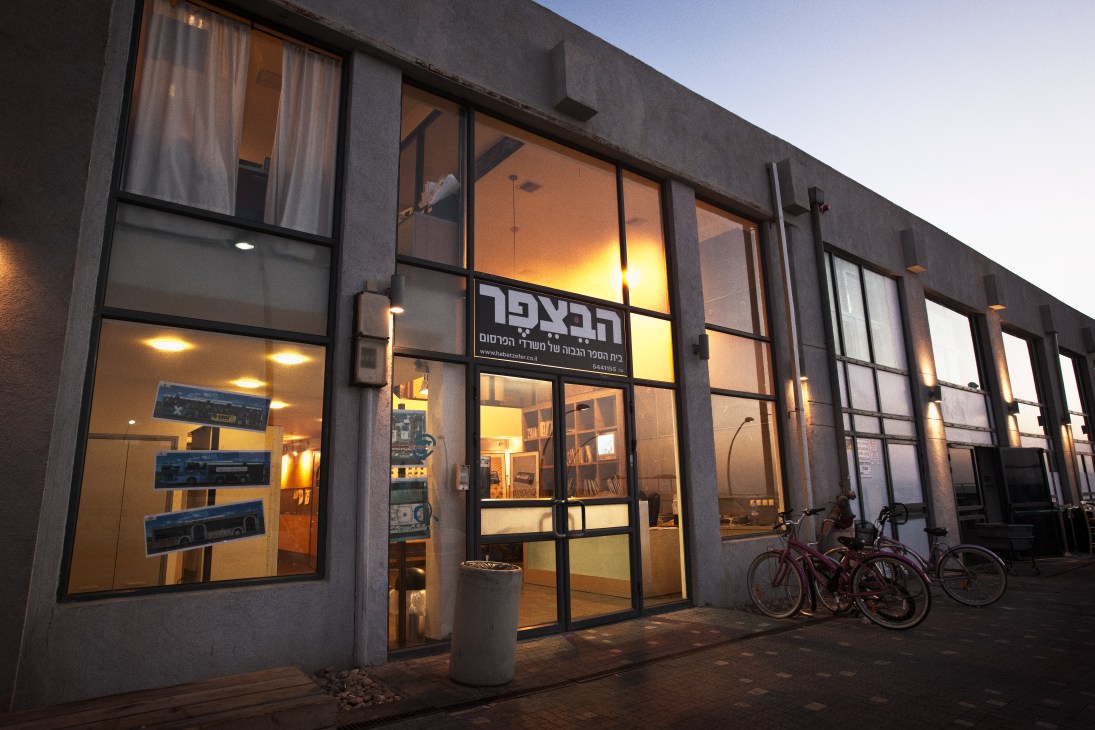 Modern glass-fronted building with Hebrew signage lit up at dusk, bicycles parked outside.