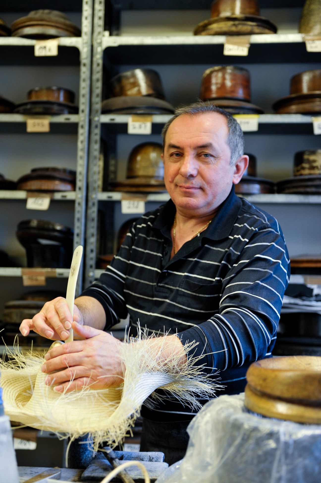 Hatter Grujica Petrovich working with straw material in the Mühlbauer atelier, with wooden hat moulds displayed on shelves behind him.