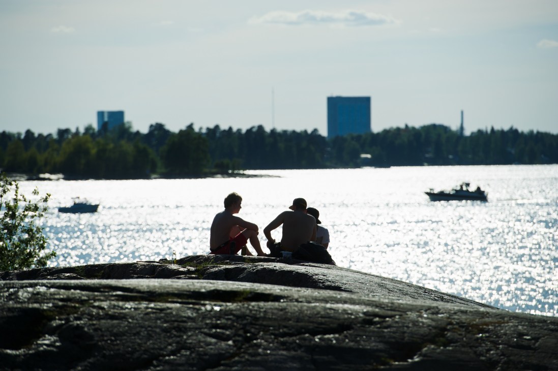 People sitting on a rocky shore overlooking a lake with Helsinki's skyline and forest in the background.