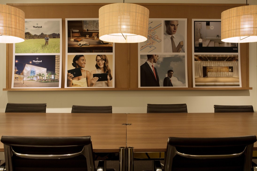 Modern boardroom with wooden table, black chairs, and wall display of framed Monocle magazine covers and photographs under pendant lighting.