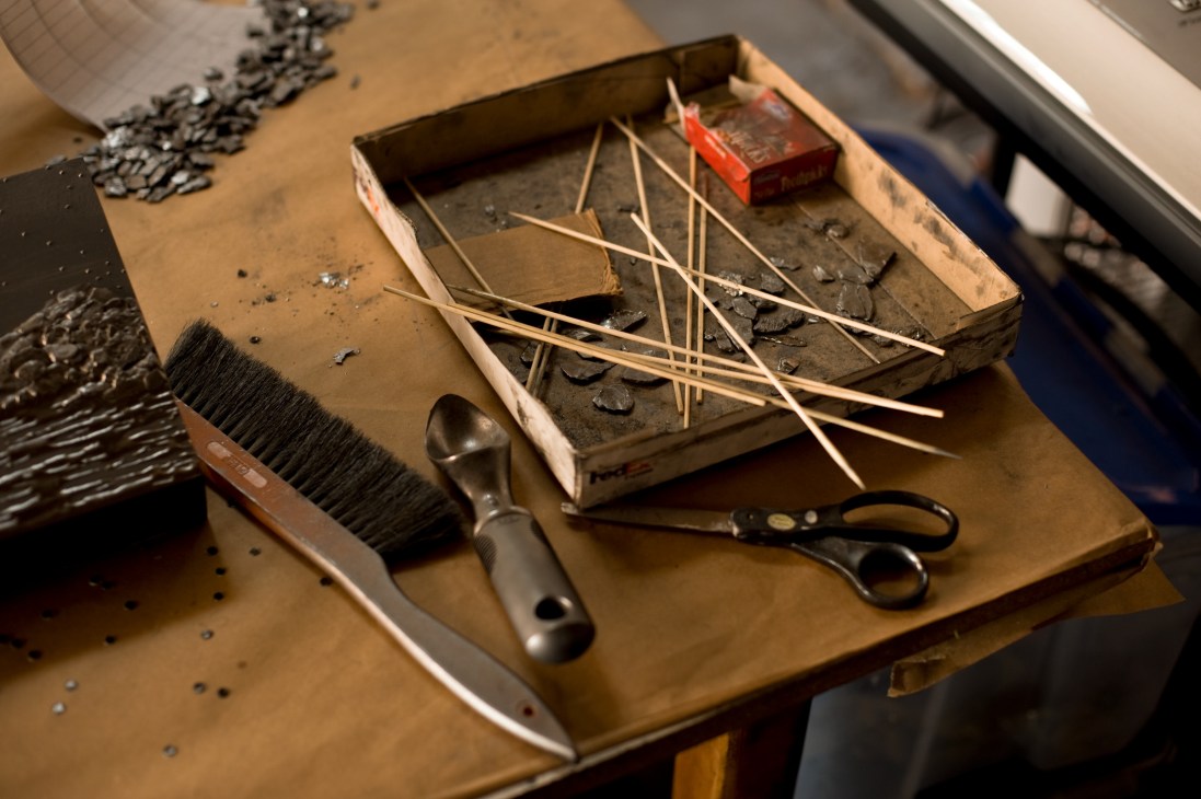 Artist's tools including brushes, scissors, and wooden sticks arranged on a tray covered in graphite dust.