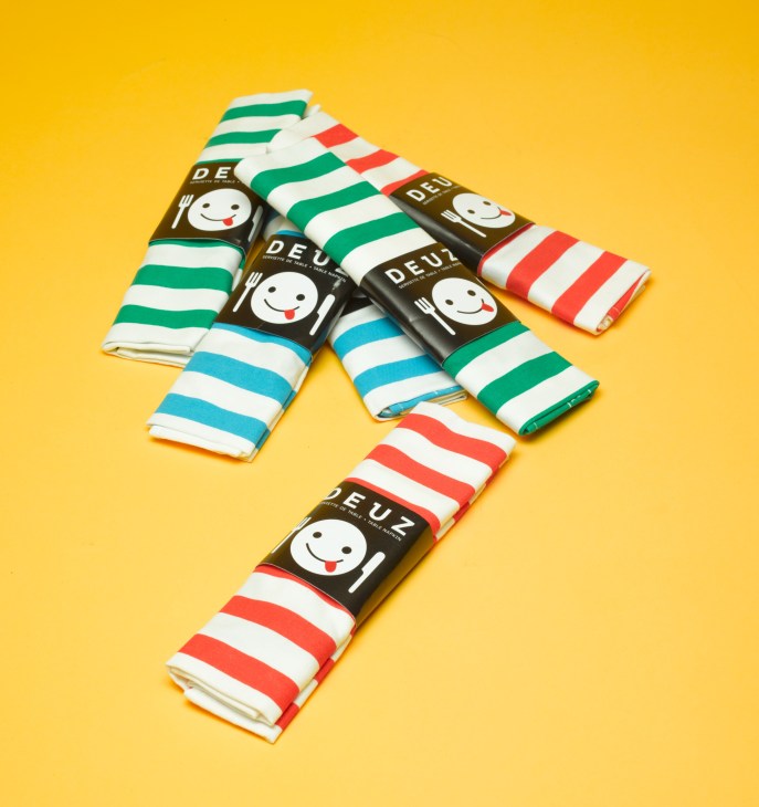 Deuz stripy napkin-aprons in green, blue, and red stripes with branded packaging on yellow background.
