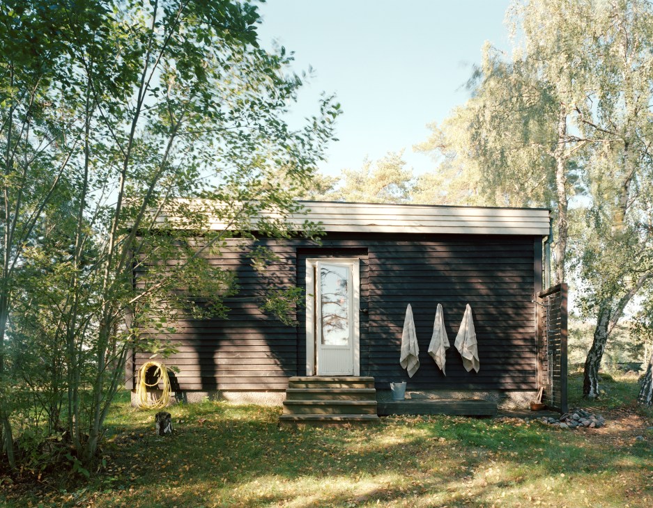 Dark wooden exterior cabin with outdoor shower, towels hanging on wall, surrounded by birch and pine trees.