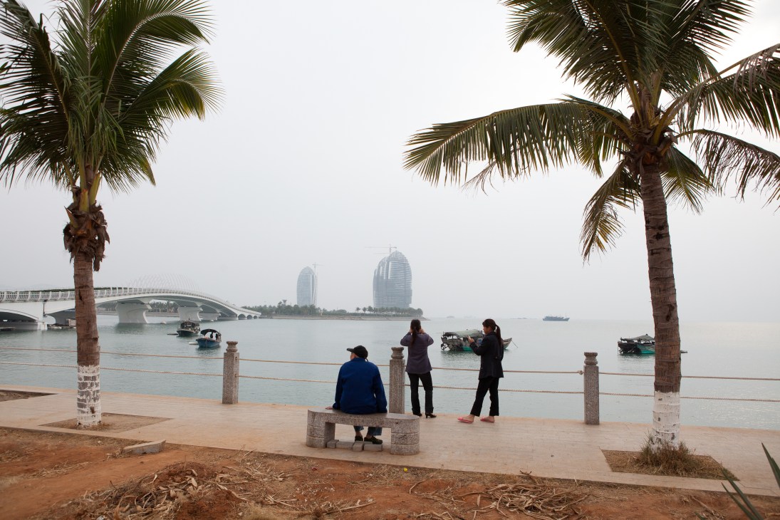 People standing on a waterfront promenade with palm trees, facing sailboats and modern buildings reflected in calm harbor water.