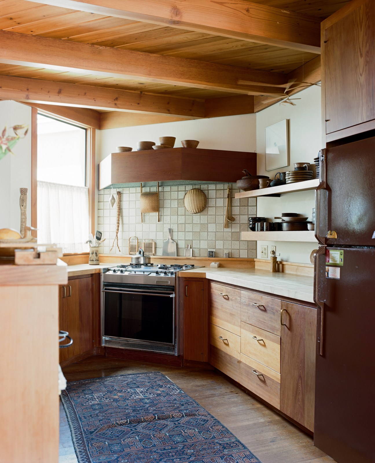 Mid-century kitchen with wooden cabinetry, angled ceiling beams, tile backsplash, and unfinished wooden bowls by Bob Stocksdale displayed on open shelving.