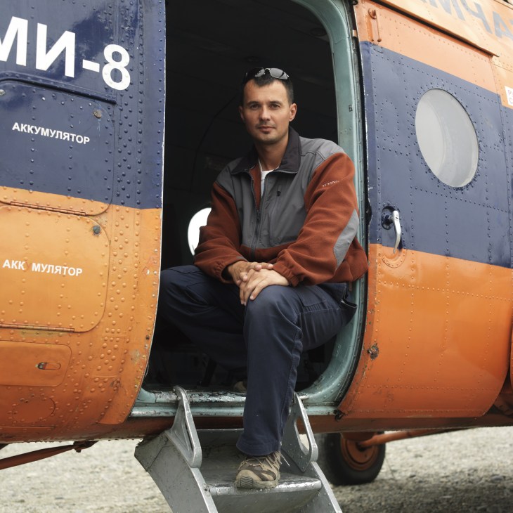 Pavel, a helicopter pilot, sits in the doorway of an orange and blue Mi-8 helicopter in Kamchatka.