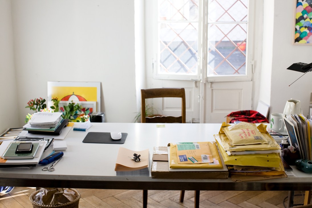 Benjamin Sommerhalder's workspace desk cluttered with papers, envelopes, books, and publishing materials in a bright room with large windows.