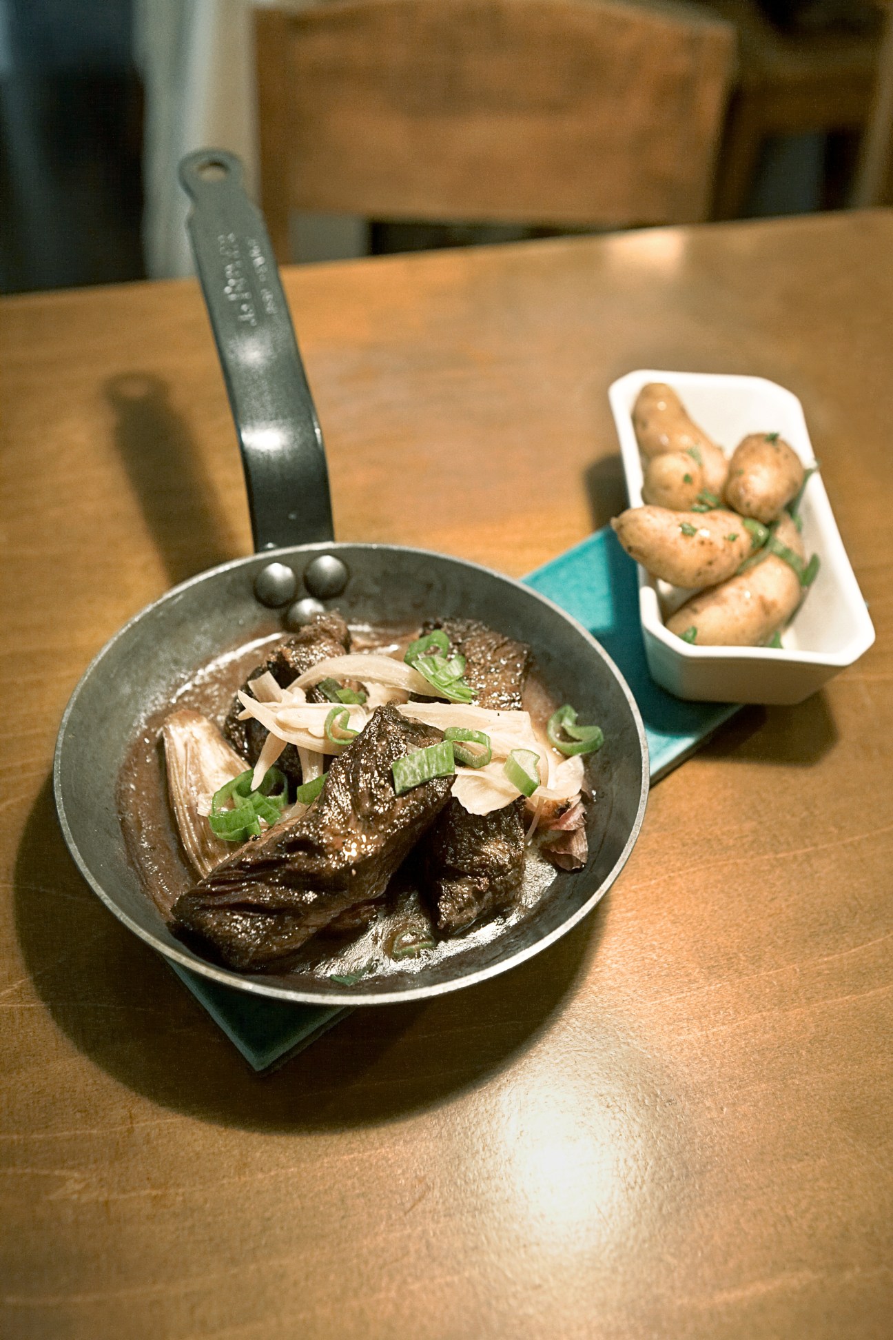 Onglet steak in a pan garnished with green onions and served with roasted potatoes.
